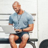 Fitnesstrainer sitzt auf Bank und arbeitet am Laptop - Dienstplan Fitnessstudio