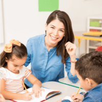 Kindergärtnerin sitzt mit Kindern am Tisch und lächelt in die Kamera - Dienstplan Kita 