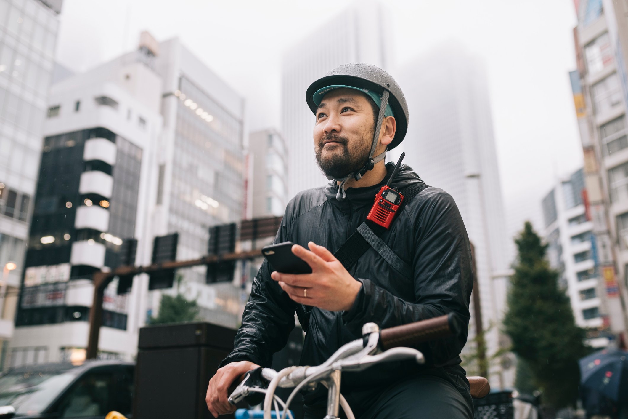 Ein freundlicher Lieferant auf einem Fahrrad in einer Großstadt. Er hat einen Helm auf, ein rotes Walki-Talki an dem Gurt seiner Umhängetasche und hält ein Smartphone in seiner Hand.