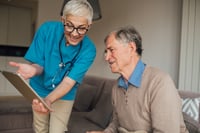 Eine medizinische Mitarbeiterin mit einem Stethoskop um den Hals lächelt freundlich, während sie einem älteren Herren etwas zeigt und erklärt.