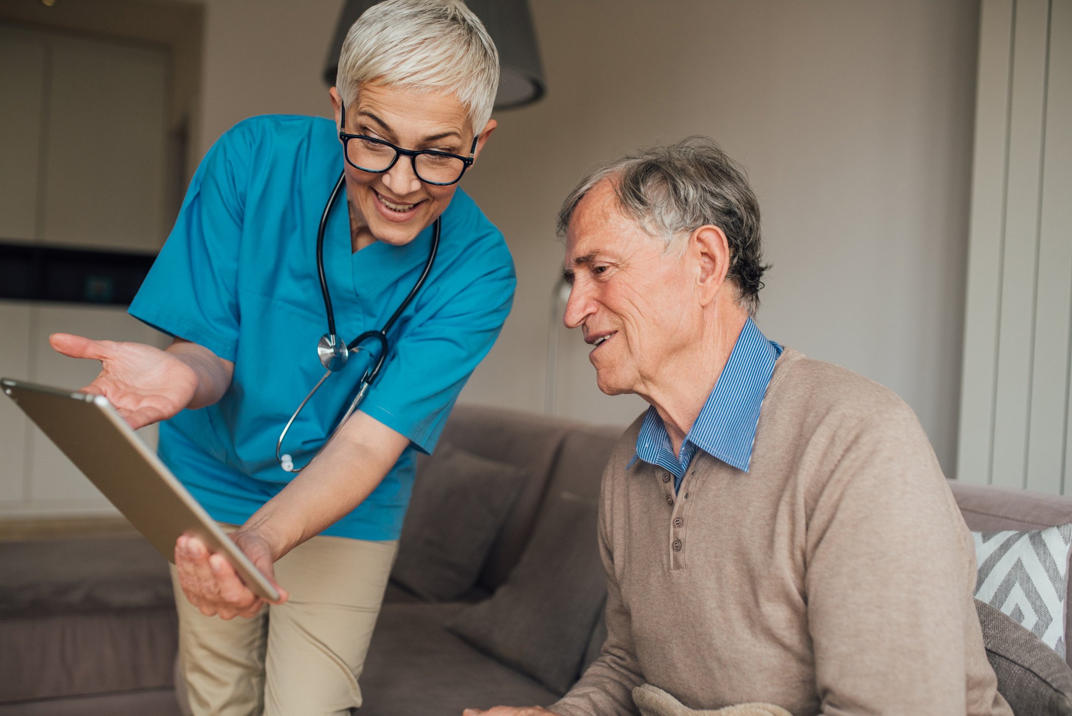 Eine medizinische Mitarbeiterin mit einem Stethoskop erklärt einem Senioren freundlich etwas auf einem Ipad.