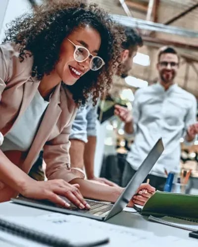 Lachende Kollegin am Laptop, im Hintergrund ein Kollege im modernen Büro